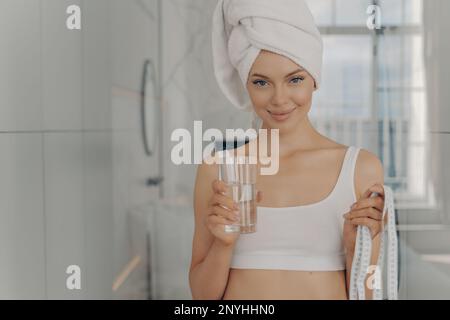 Ritratto di bella giovane femmina con pelle fresca incandescente, figura calzata e volto sorridente, tenendo un bicchiere di acqua minerale in una mano e misurare il nastro i Foto Stock