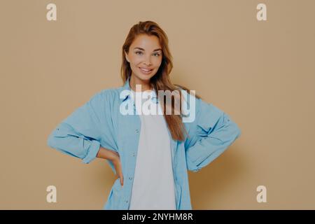 Auto sicuro sorridente modello bello mantiene le mani sulla vita, indossa una lunga camicia blu su una maglietta bianca, guarda direttamente la macchina fotografica, esprime la sua destra, isolat Foto Stock