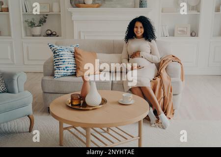 Affascinante afro americana donna incinta seduta sul divano con le mani sul suo ventre, in attesa di bambino, futura mamma guardando con gioia la macchina fotografica mentre rilassante l Foto Stock
