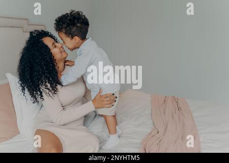 Gara mista felice donna madre incinta abbracciare adorabile bambino ragazzo mentre si siede rilassandosi a letto in camera da letto a casa. Dolci momenti di maternità di madre Foto Stock