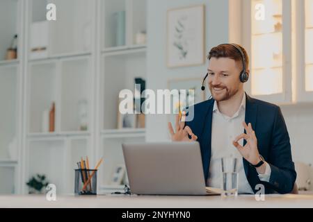 Barbuto uomo d'affari molto felice con il risultato di negoziati online mentre si utilizza il laptop e la cuffia, felice lavoratore di ufficio maschile mostrando ok segno con entrambi h Foto Stock