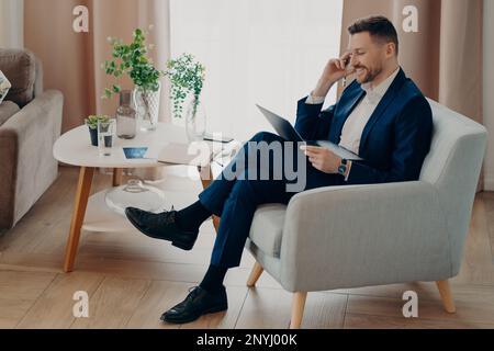 Il colpo indoor del lavoratore corporativo maschile vestito in abiti formali guarda gladfully il computer portatile tiene la penna comunica le pose online in poltrona agai Foto Stock