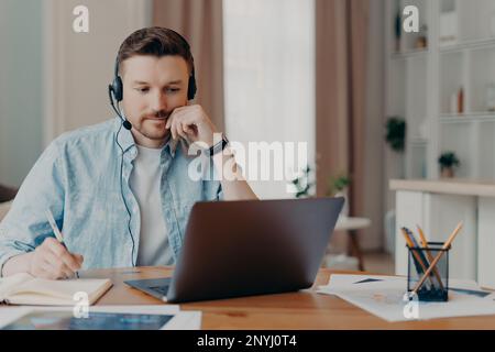 L'uomo intelligente freelance ascolta le informazioni importanti on-line fa le note necessarie in diario lavori sul laptop a casa pose sul desktop indossa jeans camicia pose ove Foto Stock