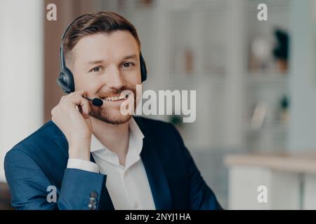 Soddisfatto allegro bearded lavoratore di ufficio maschile in tuta consulenza clienti mentre si siede e utilizzando cuffie a casa o in ufficio, concentrarsi selettivamente sul sorriso Foto Stock