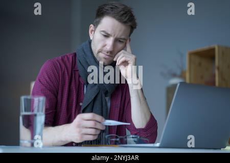 Malsano aspetto maschile freelancer sensazione di malessere e freddo, indossando calda sciarpa maglia intorno al collo, guardando sul termometro digitale nel suo controllo mano Foto Stock