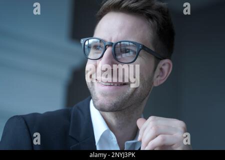 Primo piano ritratto di positivo giovane imprenditore maschile in occhiali e abbigliamento formale guardando da parte con piacevole sorriso distratto dal lavoro remoto su comp Foto Stock