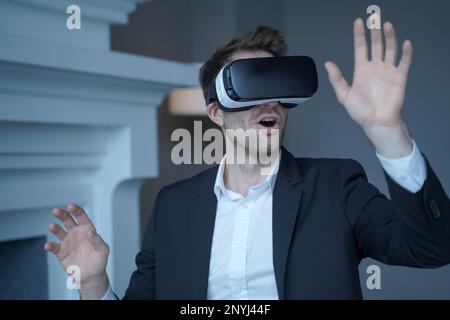 Uomo imprenditore in abito formale seduto in ufficio a casa durante il test di occhiali VR, ha colpito freddamente l'uomo d'affari che utilizza la tecnologia 3D per la prima volta con Foto Stock