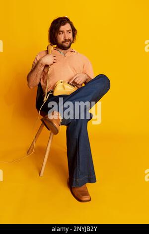 Emotivo uomo barbuto, hippie in stile vintage jeans svasato seduta e parlare su telefono retrò su sfondo giallo. Concetto di moda, emozioni Foto Stock
