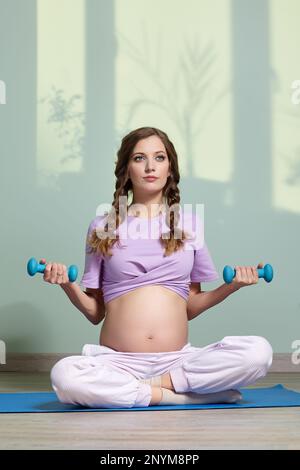 Una donna incinta con manubri in mano siede a gambe incrociate, meditando e praticando yoga per il fitness fisico durante la gravidanza. Sul retro Foto Stock