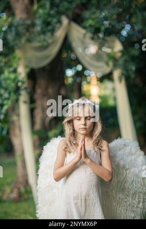 Bambina a piedi nudi in abito bianco con ali d'angelo e corona di fiori che prega in giardino con un'arcata di tessuto sognante appesa all'albero Foto Stock