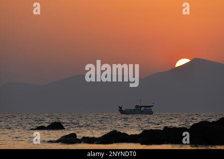 una solitaria barca da pesca in mare mentre il sole tramonta dietro le montagne Foto Stock