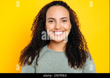 Foto ravvicinata di una bella giovane donna da corsa mista sorridente, con capelli ricci, vestita con una maglietta sportiva, in piedi su sfondo giallo isolato, guardando la macchina fotografica, con un sorriso tondo Foto Stock