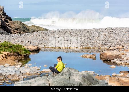 Giovane ragazzo in maglione giallo appoggiato su una roccia su un lungomare roccioso a Los Molinos, Fuerteventura. L'onda si blocca sullo sfondo. Foto Stock