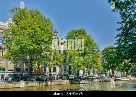 Amsterdam, Paesi Bassi - 10 aprile 2021: Alcuni edifici e alberi sul lato di un fiume con barche parcheggiate lungo di esso, di fronte a una fila di case Foto Stock