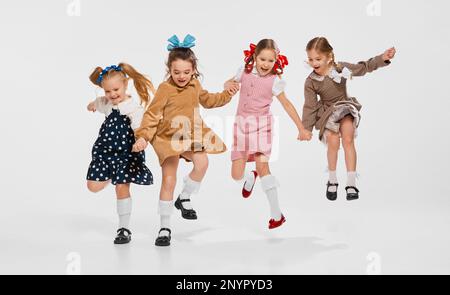 Quattro belle bambine, bambini in abiti retrò alla moda in posa, saltando contro sfondo grigio studio. Concetto di infanzia Foto Stock