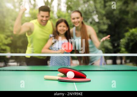 Famiglia vicino al tavolo da ping pong nel parco, focalizzazione su racchette e palla Foto Stock