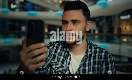 Sorridente arabo caucasico maschio blogger uomo d'affari guardare il telefono cellulare fotocamera parlare videochiamata nel centro commerciale felice giovane uomo chattando online Foto Stock