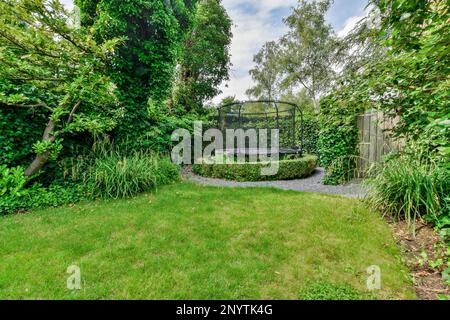 un giardino con erba verde e cespugli sullo sfondo, vi è una panca circolare in pietra circondata da una vegetazione lussureggiante Foto Stock
