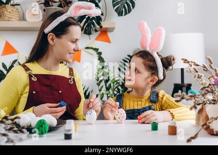 Felice madre giovane con sorridente bambina per tingere e decorare le uova con pitture per le vacanze di Pasqua, mentre seduti insieme a un tavolo bianco in Foto Stock