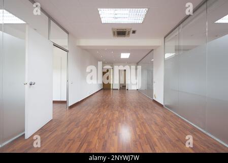 Uffici vuoti con soffitti tecnici, pareti bianche, pavimenti in legno rossiccio e divisori in vetro temprato con porte trasparenti Foto Stock