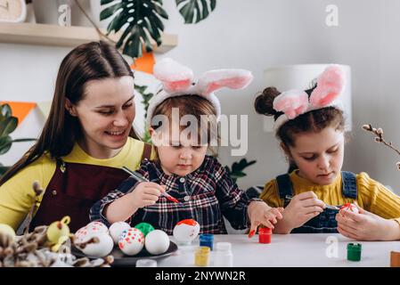 Felice giovane mamma caucasica e due figlie che decorano le uova con le pitture per le vacanze di Pasqua mentre si siedono insieme al tavolo bianco. Famiglia divertente Foto Stock