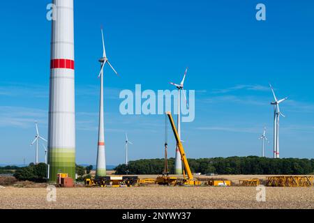 Una gru da costruzione gialla sta assemblando una struttura metallica in un campo davanti ai mulini a vento. Foto Stock