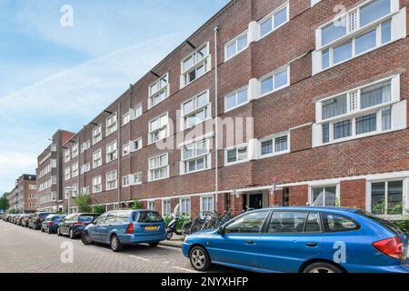 Amsterdam, Paesi Bassi - 10 aprile 2021: Auto parcheggiate sul lato di una strada di fronte a un edificio in mattoni con finestre bianche e pareti in mattoni rossi Foto Stock