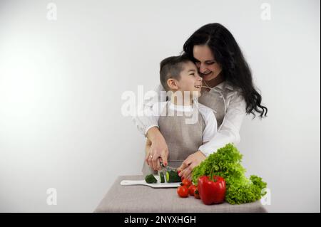 rapporti di famiglia padre al lavoro mamma e figlio cuoco cena a casa mamma insegna al figlio a tagliare le verdure per insalata ragazzo tagli cetriolo mamma abbracciato e lo aiuta in modo da non danneggiare la sua mano attitudine di cura Foto Stock