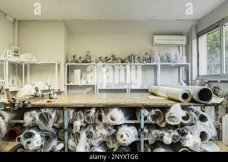 Un tavolo in legno di una piccola officina di lampade con tutti i tipi di materiali per la sua costruzione su mensole in metallo e sotto il tavolo Foto Stock