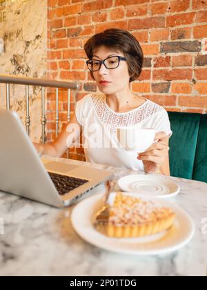 Donna lavora a distanza in un caffè con pareti di mattoni rossi. Femmina nei tipi degli occhiali sulla tastiera del laptop, beve il caffè e mangia una crostata. Centro di lavoro Foto Stock