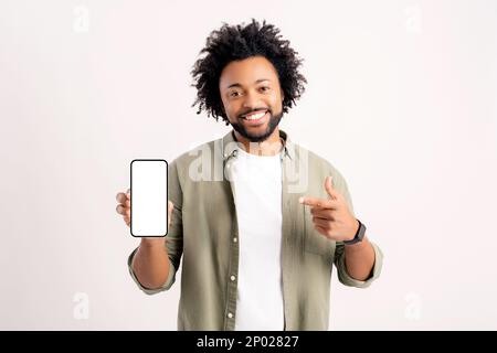 Attraente afro-americano 30s uomo che punta il dito contro lo smartphone con schermo vuoto in piedi isolato su bianco, positivo riccio ragazzo con un sorriso toothy che punta il dito contro il dispositivo telefonico, mock up Foto Stock