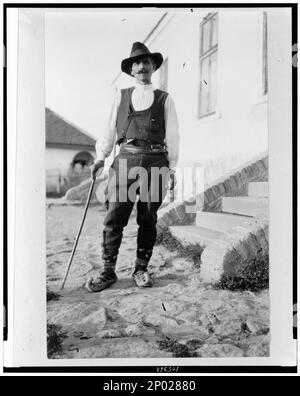 Uomo serbo che indossa cappello, giubbotto, pantaloni con cintura e scarpe a punta con canna fuori dall'edificio in Jugoslavia. Frank and Frances Carpenter Collection, No. 963, Serbi, Abbigliamento e vestito, Yugoslavia, 1880-1930. Foto Stock