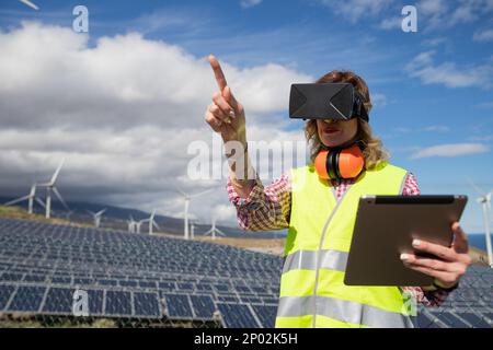 Un ingegnere femminile presso la fattoria solare lavora utilizzando le cuffie per la realtà virtuale e il tablet. Foto Stock