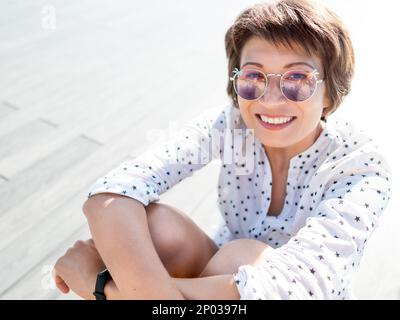 Donna con i capelli corti e le pose dei faretti in occhiali da sole colorati. Donna sorridente in una scena di legno aperto del parco urbano. Atmosfera estiva. Emozioni sincere. Foto Stock