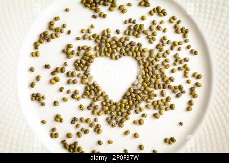 Fagioli in una forma del cuore isolato su una piastra bianca. Materie green gram semole ingrediente alimentare dello sfondo. Amore per il concetto di cucina a vista Foto Stock