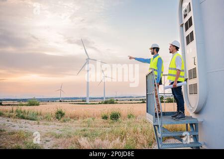 Due ingegneri o tecnici di mulino a vento che progettano una costruzione che indica dove la turbina nuova sarà messa. Un paio di lavoratori delle energie rinnovabili che indossano elmetti o casco e giubbotti riflettenti. Concetto di industria sostenibile. Foto di alta qualità Foto Stock
