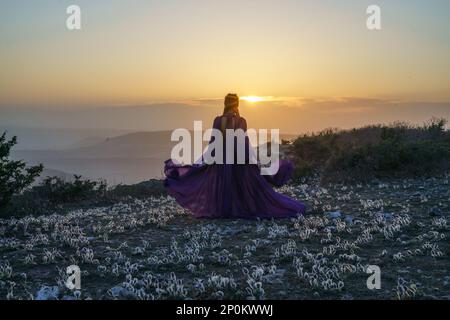 Tramonto viola abito donna montagne. Ascesa del mistico. tramonto sulle nuvole con una ragazza in un lungo vestito viola. Nel prato c'è un prato Foto Stock