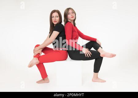 Donna mamma e figlia seduta con le spalle l'una all'altra su cubo su sfondo bianco. Entrambe le donne hanno attraversato le gambe e guardando seriamente Foto Stock