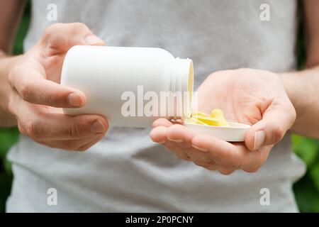 Uomo in una t-shirt grigia che versa le pillole da un vaso di plastica bianca. Primo piano, copia spazio, spazio per etichetta o branding. Foto Stock