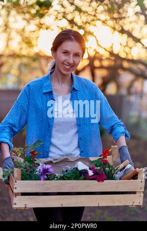 Donna giardiniere in abbigliamento casual tiene scatola di legno con vasi di fiori. Casa giardinaggio accogliente. Piccola impresa femminile, negozio di fiori, flowerpot, freelance, Foto Stock