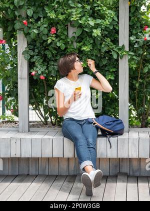 La donna sta anniettendo le rose del trellis. Bella donna è seduta con smartphone su una panca di legno nel parco pubblico. Atmosfera estiva. Foto Stock