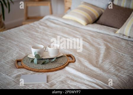 Sul copriletto di lino si trova un vassoio rotondo di legno, su di esso ci sono due tazze vuote di ceramica, eucalipto verde. Mattina elegante e alla moda. Textural, Scandinavian R Foto Stock