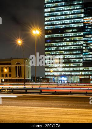 luce proveniente dal passaggio di auto sulla strada innevata della città e dalle luminose finestre di un alto edificio in vetro sullo sfondo nella notte d'inverno Foto Stock