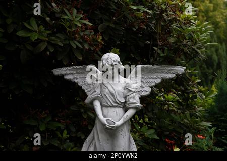 Una maestosa statua d'angelo si trova in una zona erbosa, con alberi alti che forniscono uno sfondo sereno Foto Stock