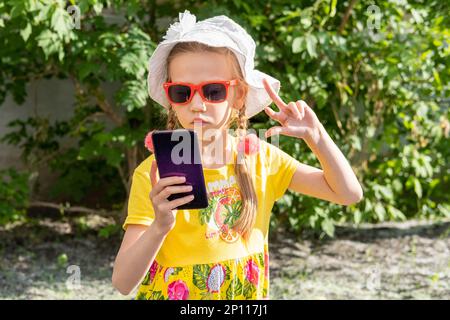 Ritratto di bambina che prende un selfie da smartphone nel parco estivo. Una ragazza allegra in un vestito giallo, cappello bianco e occhiali da sole prende un Foto Stock