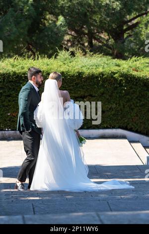 La sposa in un lungo vestito bianco si erge sulle scale del parco accanto allo sposo in una giornata estiva. Russia, Adler, 10 maggio 2022 Foto Stock