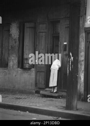 Vista dall'altra parte della strada di una donna in piedi in una porta nel quartiere francese, New Orleans, tra le 1920:1926 e le 17:00. Foto Stock