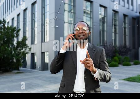 Un uomo d'affari afroamericano di successo è in piedi vicino ad un centro ufficio in un vestito e occhiali e parlando al telefono. Foto Stock
