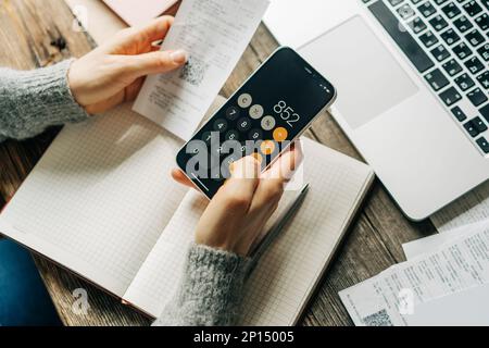 Donna irriconoscibile lavora alla sua scrivania con bollette utilizzando una calcolatrice sul suo telefono cellulare. Foto Stock