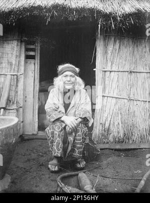 Ainu donna seduto di fronte alla porta di una capanna, 1908. Foto Stock